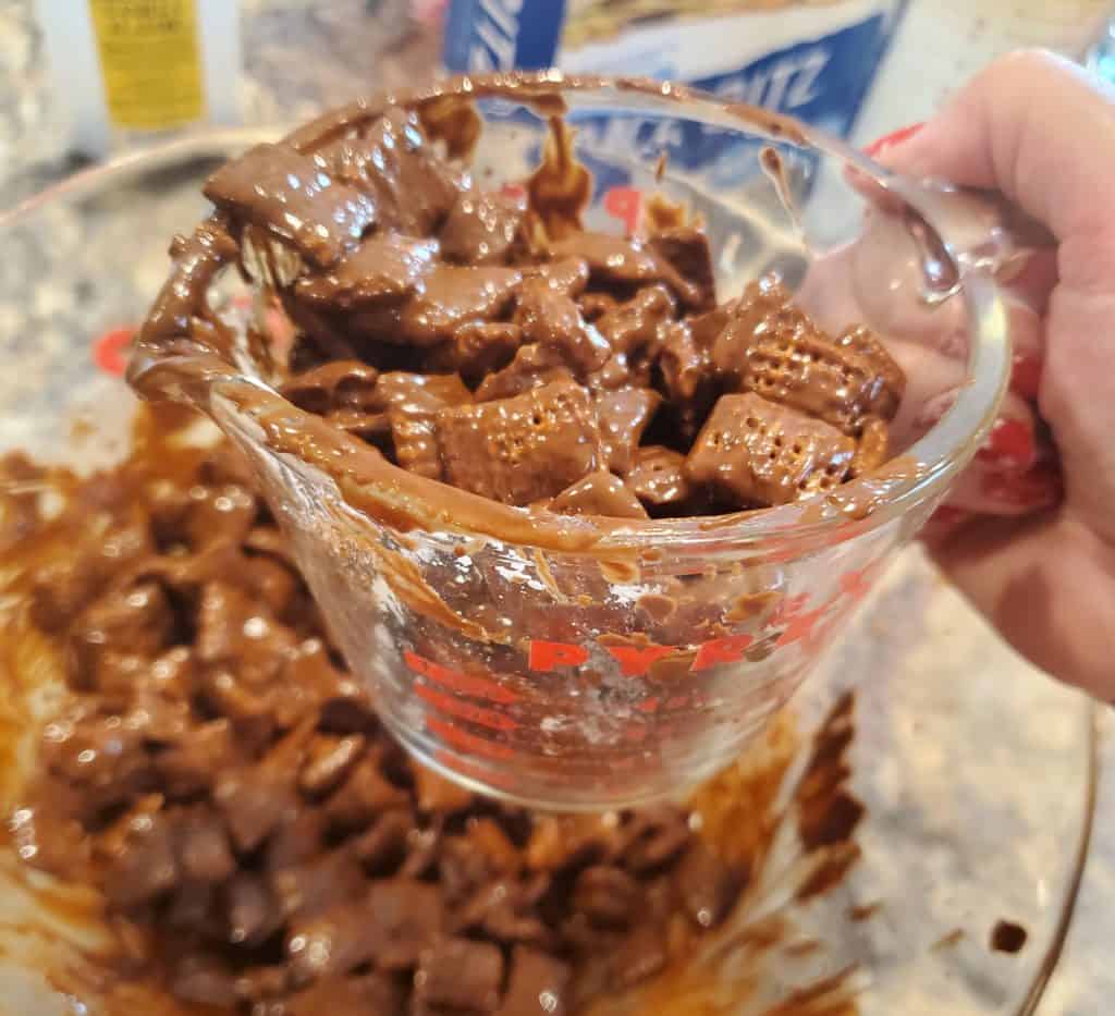 Scooping out the cereal coated in the peanut butter and chocolate mixture of this puppy chow recipe with a measuring cup