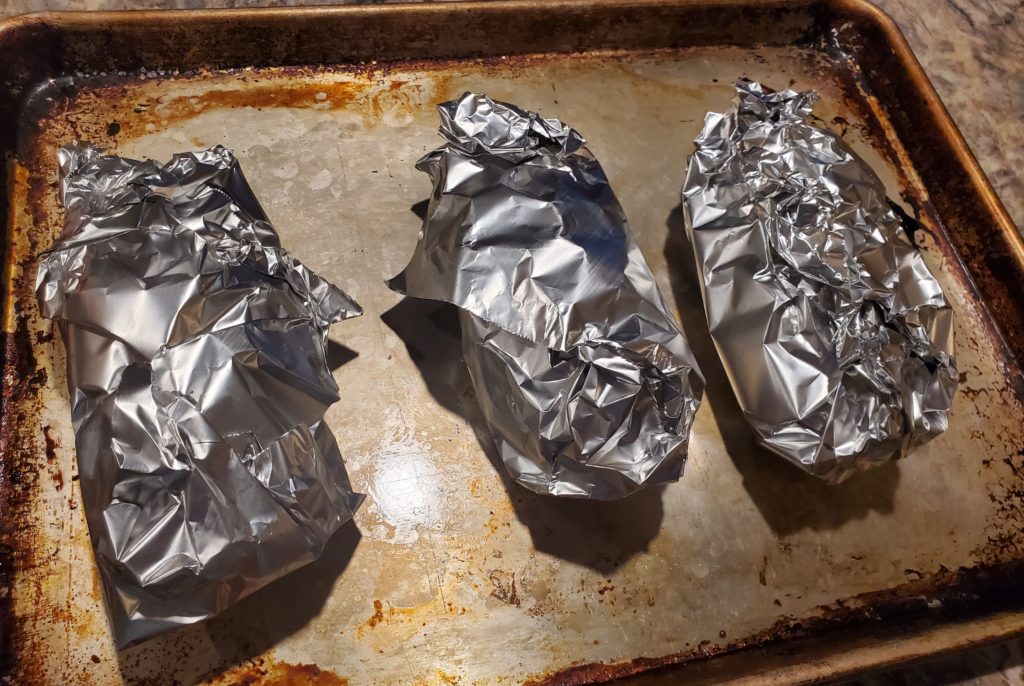 Three potatoes wrapped in foil on a baking sheet and ready to go into the hot oven