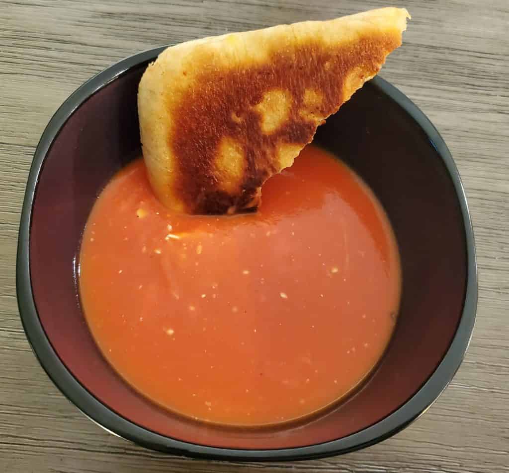 A bowl of creamy tomato soup with half a ciabatta grilled cheese sandwich garnishing it