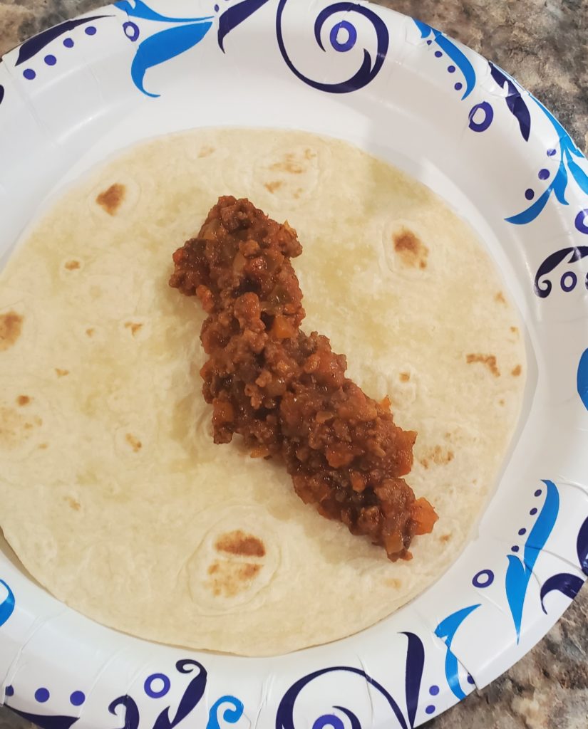 A beef enchilada on a paper plate ready to be rolled