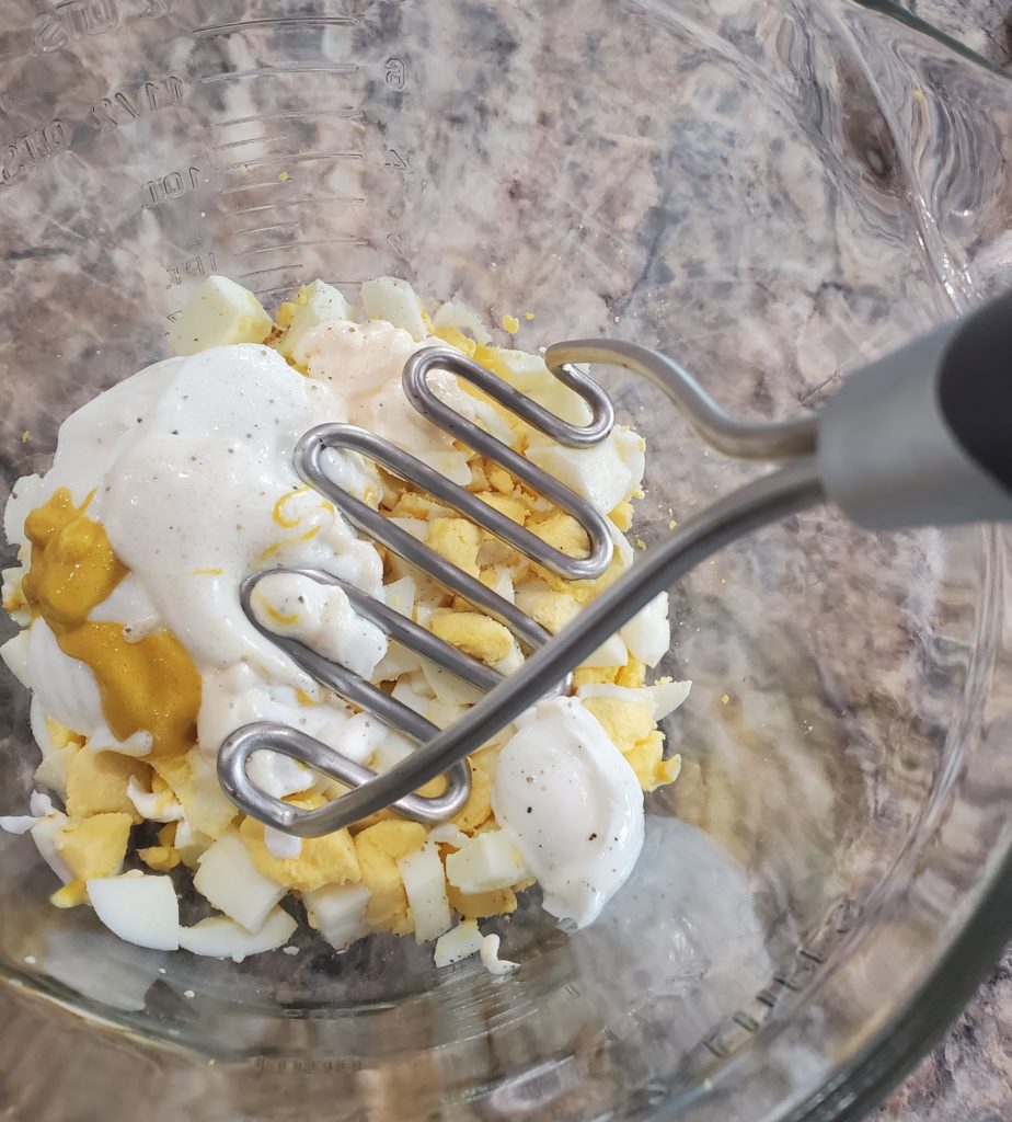 A potato masher being used to mash all the egg salad ingredients together